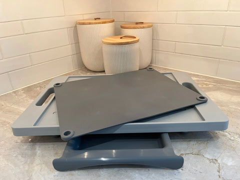 The Karving King Dripless Cutting Board 2 in 1 System, featuring three nested gray boards, rests on a marble countertop with white textured canisters and wooden lids set against a white tile backsplash in the kitchen.