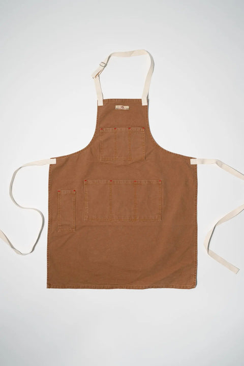 The Common Apron Canvas by Marc Nelson, a brown canvas apron with adjustable straps, three front pockets, one side pocket, and beige neck and waist ties, is laid flat on a white background.