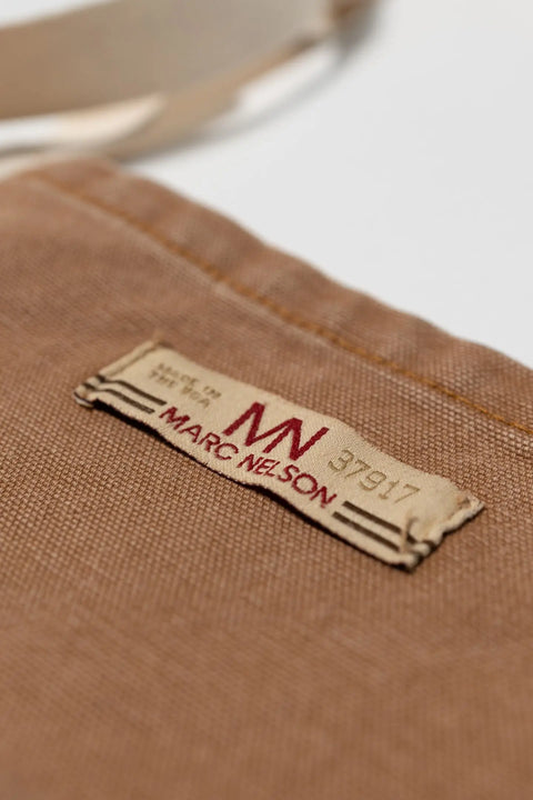 A close-up of The Common Apron Canvas by Marc Nelson shows its brown fabric, multiple pockets, and a slightly frayed label displaying MARC NELSON in red text with black numbers 37917.