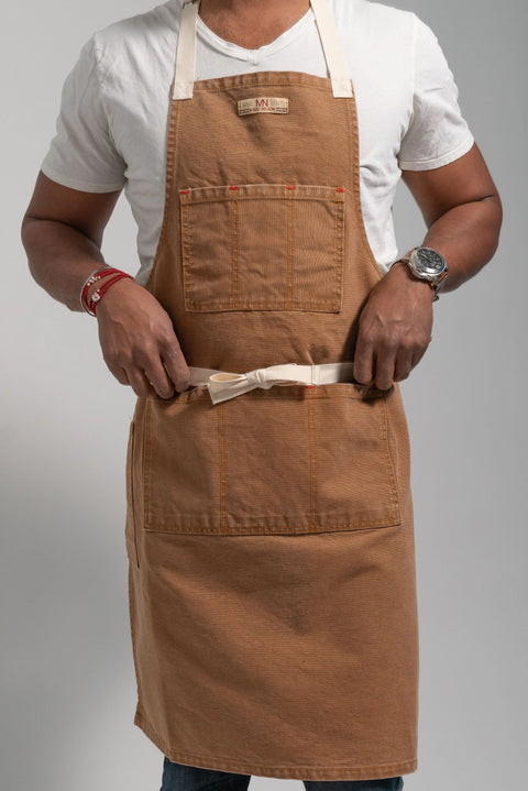 A person wearing Marc Nelsons The Common Apron Canvas—a brown apron with multiple pockets and a beige adjustable waist strap—over a white t-shirt is tying the strap; bracelets and a wristwatch accent, with a plain light background.
