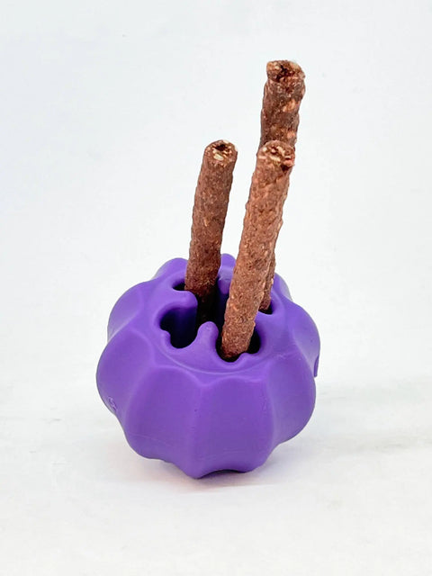 The SodaPup Cross Bones Chew Toy and Treat Pocket, a purple pumpkin-shaped toy, holds three brown stick-shaped dog treats upright in its holes against a plain white background.