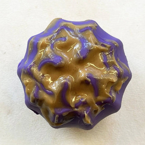 The SodaPup Cross Bones Chew Toy and Treat Pocket, a round chew toy coated in purple with a drizzle of light brown peanut butter, sits on a white textured surface.
