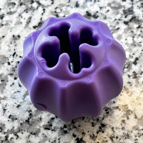 The SodaPup Cross Bones Chew Toy and Treat Pocket, a round purple rubber toy with wavy grooves, sits on a speckled granite countertop.