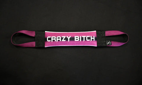 A pink and white Crazy Bitch Fire Hose Training Tug by Bulletproof Pet Products Inc, with black handles on each end, lies flat on a black surface. CRAZY BITCH is printed in bold white letters across the center.