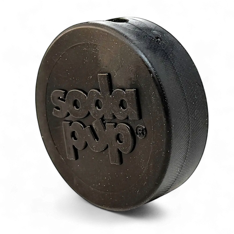 The SodaPup Hockey Puck Durable PUP-X Rubber Treat Holder and Chew Toy, a black round toy with SodaPup embossed on it, is displayed against a solid black background.