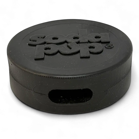 A black SodaPup Hockey Puck Durable PUP-X Rubber Treat Holder and Chew Toy, featuring a rectangular slot for bully sticks and soda pup embossed on top, sits against a black background.