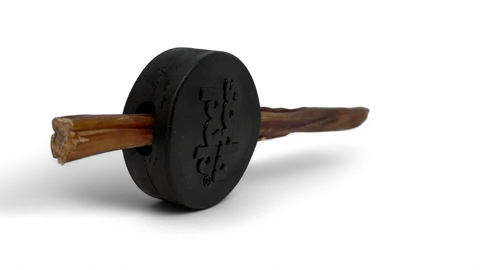 The SodaPup Hockey Puck Durable PUP-X Rubber Treat Holder and Chew Toy is a round, black dog toy with an embossed logo, holding a brown chew stick in its center, shown on a black background.