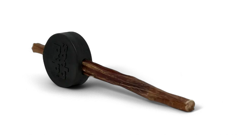 A chewy brown treat stick is inserted into the SodaPup Hockey Puck Durable PUP-X Rubber Treat Holder and Chew Toy, both featuring embossed text and shown isolated against a black background.