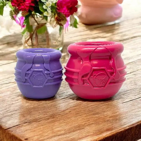 Two SodaPup Honey Pot Durable PUP-X Rubber ECup Treat Dispenser & Enrichment Toys, one purple and one pink, sit on a wooden table with flowers in the background. Each features a whimsical honey pot design with a hexagon emblem on the front.