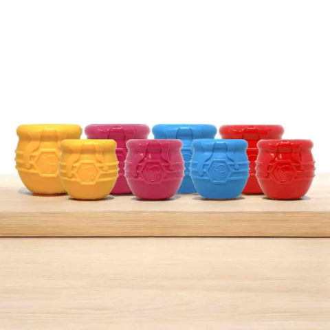 Eight small Honey Pot Durable PUP-X Rubber ECup Treat Dispenser & Enrichment Toys by SodaPup—three yellow, two pink, two blue, one red—are arranged in two rows on wood against a white background.