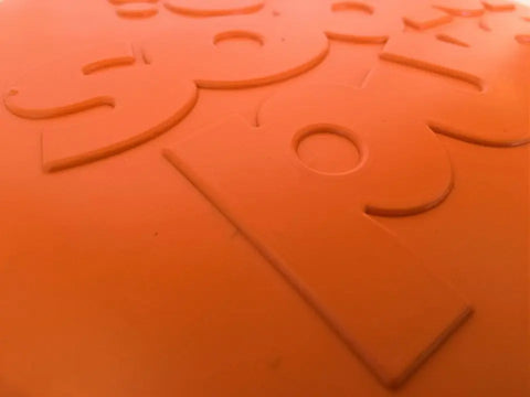 Close-up of raised, embossed text and numbers on the orange SodaPup Bottle Top Flyer Durable Rubber Retrieving Frisbee. Large letters and shapes are highlighted, though some of the text is partially out of frame.