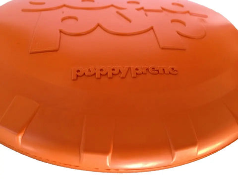 Close-up of the orange SodaPup Bottle Top Flyer Durable Rubber Retrieving Frisbee with puppyprene embossed in the center and part of a larger logo above it.
