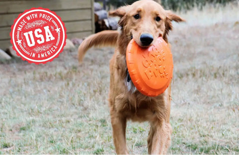A golden retriever runs on grass with a SodaPup Bottle Top Flyer Durable Rubber Retrieving Frisbee in bright orange in its mouth. A red “Made With Pride in America, USA” badge appears in the upper left corner of the image.