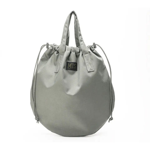 The MIS UK Helmet Bag - Foliage is a round, light gray Cordura Nylon bag with two handles, a black MIS label on the front, and adjustable drawstring sides, shown against a white background.