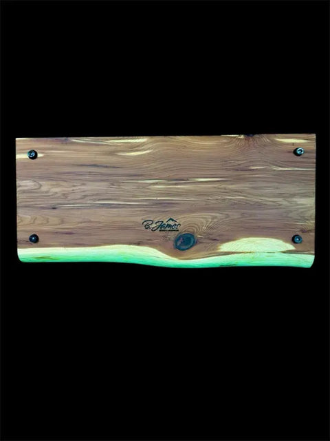 The Eastern Red Cedar Live Edge Charcuterie Board by B. James Design & Woodwork features natural wood grain, a knot, a mountain logo in the center, rounded edges, and four corner screws—all set against a black background.