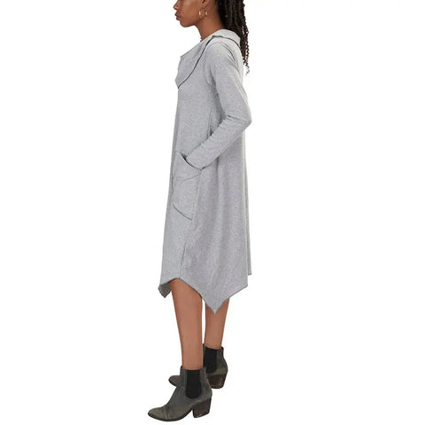 A person wears the Spiritex | Sustainable Apparel 50/50 Long Sleeve Colette Drape Dress, featuring an asymmetrical hem and A-line drape with side pockets, styled with black ankle boots, standing in profile against a white background.