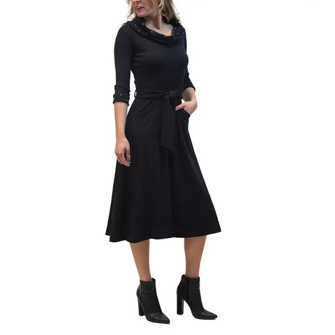 A woman wears the French Broad Dress by Spiritex | Sustainable Apparel, a black organic cotton, long-sleeve knee-length dress with cowl neck and waist tie, styled with black heeled ankle boots; one hand in her pocket, hair loose.