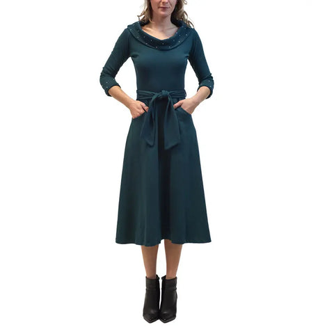 A woman wears the Spiritex | Sustainable Apparel French Broad Dress—a teal, 50s-inspired style with long sleeves, cowl neck with small embellishments, waist tie, and pockets—paired with black ankle boots against a plain white background.