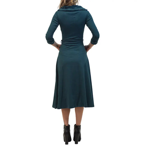 A woman stands facing away, wearing a teal French Broad Dress by Spiritex | Sustainable Apparel, featuring long sleeves, organic cotton fabric, and a knee-length hem, paired with black heeled ankle boots. Her hands rest on her hips against a white background.