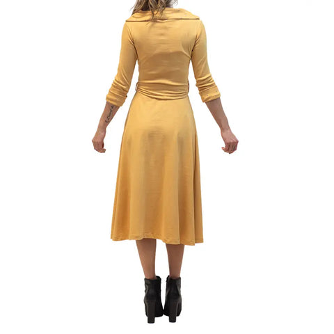 A woman faces away, wearing a long-sleeved, knee-length yellow French Broad Dress by Spiritex | Sustainable Apparel and black heeled ankle boots, set against a plain white background.