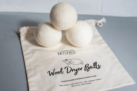 Three white wool dryer balls from American Blossom Linens’ Reusable Dryer Ball Set Natural Wool rest on a branded cloth bag with a sheep design—an eco-friendly alternative to fabric softeners for your laundry.
