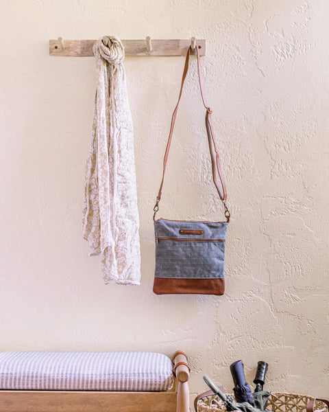 A light scarf and a blue-and-brown Ellis River Crossbody Bag by Rogue Industries hang on a wooden wall hook above a cushioned bench. Below, an umbrella rests in a basket. The textured wall is painted light cream.