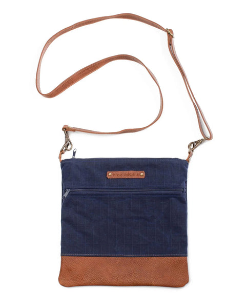 The Ellis River Crossbody Bag by Rogue Industries features a blue fabric body, brown leather bottom, long adjustable brown strap, front zip pocket, and a small brown leather patch with text on the front.