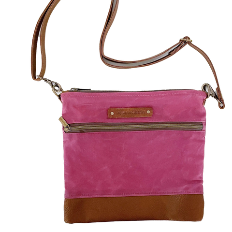 The Ellis River Crossbody Bag by Rogue Industries features a pink fabric body, brown leather bottom and strap, two zippered compartments, and a front leather patch with text. This stylish crossbody purse is shown on a white background.