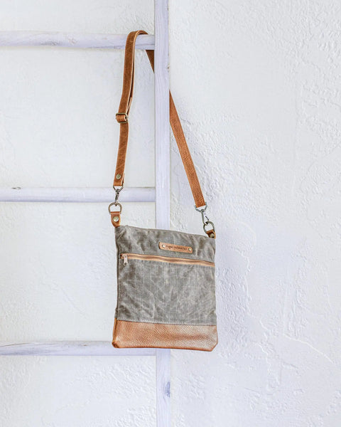 The Ellis River Crossbody Bag by Rogue Industries, featuring a gray fabric body, brown leather accents, and a front zipper pocket, hangs from a white wooden ladder against a textured white wall.