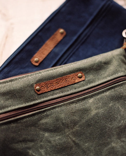 A close-up of two Ellis River Crossbody Bags by Rogue Industries, made from durable waxed canvas—one olive green, one navy—each with a brown leather label embossed “Rogue Industries” and secured with brass rivets.