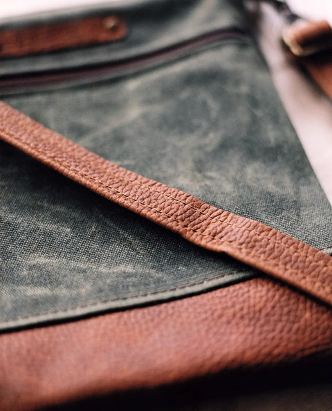 Close-up of the Ellis River Crossbody Bag by Rogue Industries, featuring green waxed canvas, a textured brown leather strap and trim, detailed stitching, and a visible zipper.