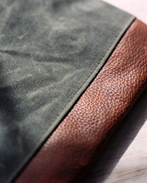 A close-up of the Rogue Industries Ellis River Crossbody Bag highlights its textured brown leather edge and green waxed canvas, displayed on a light wooden surface.