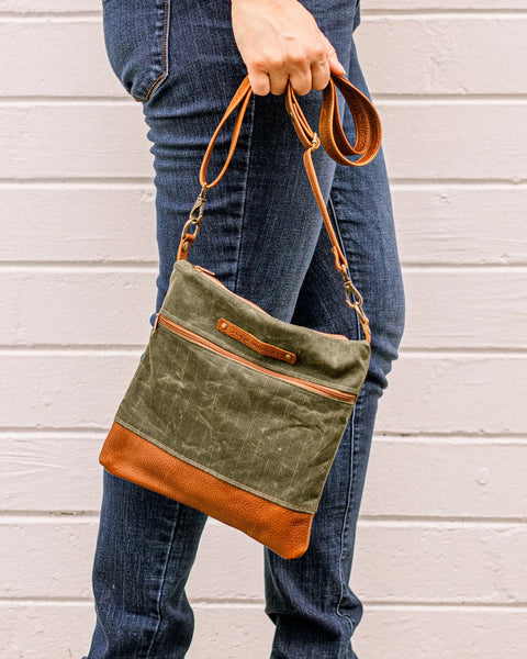 A person in blue jeans holds the Rogue Industries Ellis River Crossbody Bag—a green waxed canvas bag with brown leather accents—in front of a white wooden wall.