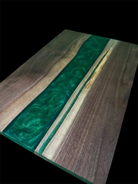 The Emerald Green Epoxy and Walnut Serving Board by B. James Design & Woodwork is handcrafted in the USA, featuring a river-like strip of glossy green epoxy between natural walnut wood grain, all set against a black background.