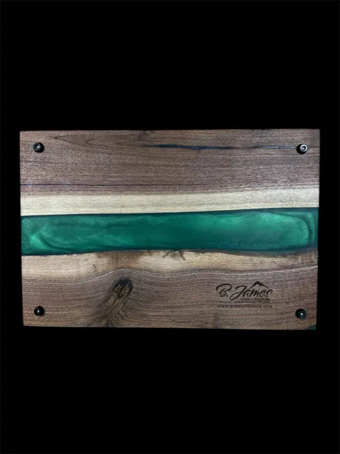 The Emerald Green Epoxy and Walnut Serving Board by B. James Design & Woodwork features a horizontal green epoxy river, four metal-corner screws, and a wood-burned logo in the bottom right. Handcrafted in the USA; shown on a black background.