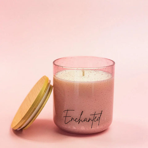 The Enchanted Scented Candle by Shanti + Tula, in pink glass with a wooden lid, fills your room with the exotic aromas of raspberry, lychee & vanilla. Handcrafted in the USA and beautifully set against a soft pink background.