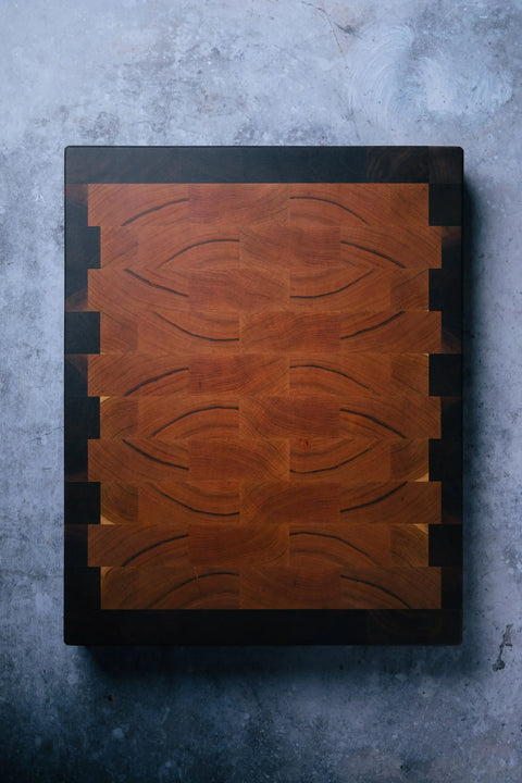 The Montana Block The Chef - End Grain Butcher Board - Cherry is a square, American-made cutting board with an intricate geometric end grain pattern in warm cherry and dark wood tones, displayed on a textured gray surface.
