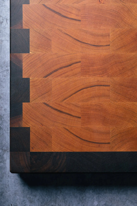 Close-up of The Chef - End Grain Butcher Board - Cherry by Montana Block, featuring interlocking dovetail joints, natural cherry wood grain, and dark-light tones, displayed on a gray textured surface.