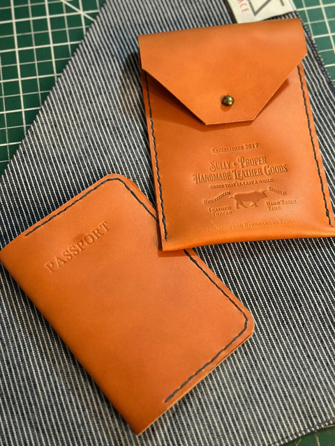 Two brown English Bridle Leather travel accessories from Sully + Proper Supply—a passport cover and a utility pouch—lie on striped fabric. Both handmade items feature visible stitching and are part of the Travel Bundle set.