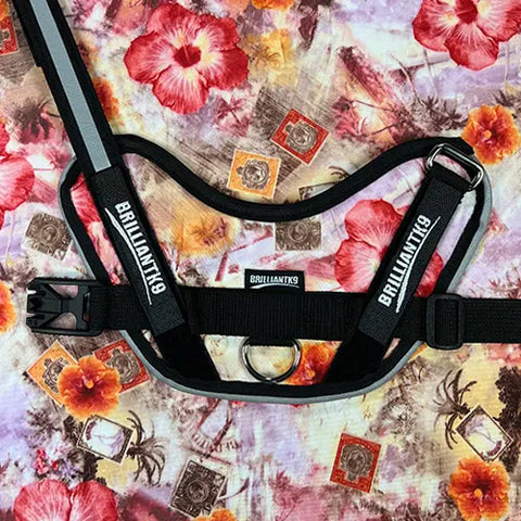 A BrilliantK9 Ergonomic Dog Harness for small dogs (10–20 lbs) by BrilliantK9 in black rests on a colorful fabric with red hibiscus flowers, palm trees, and postage stamp patterns.