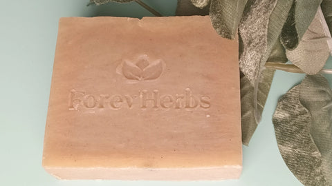 A beige square bar of Forev'Herbs Eucalyptus Lime Handmade Soap, featuring a leaf logo, sits by green velvety leaves on a light blue surface. Made with natural fragrance oils for a refreshing scent.