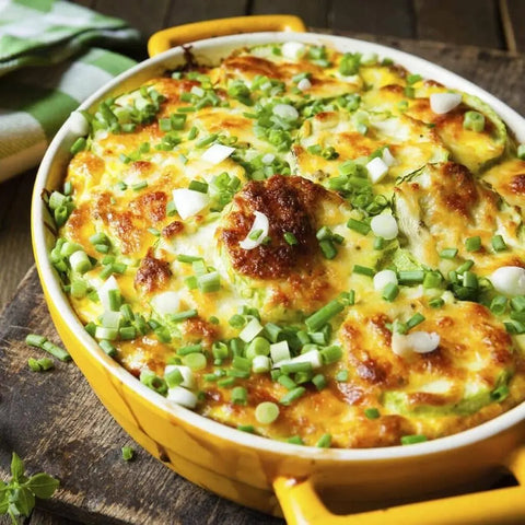 A baked casserole in a yellow dish, topped with melted cheese, green onions, herbs, and Gustus Vitae Italian and French Countryside Spice Blend Collection—3 Tins, sits on a wooden surface beside a green and white cloth.