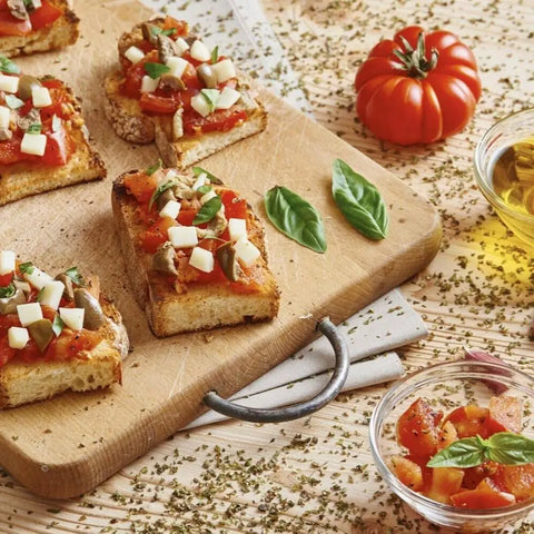 Slices of bruschetta topped with tomatoes, mozzarella, and basil are served on a wooden board. A fresh tomato, basil leaves, and a glass of olive oil highlight the artisan touch of Gustus Vitae’s Italian and French Countryside Spice Blend Collection - 3 Tins.
