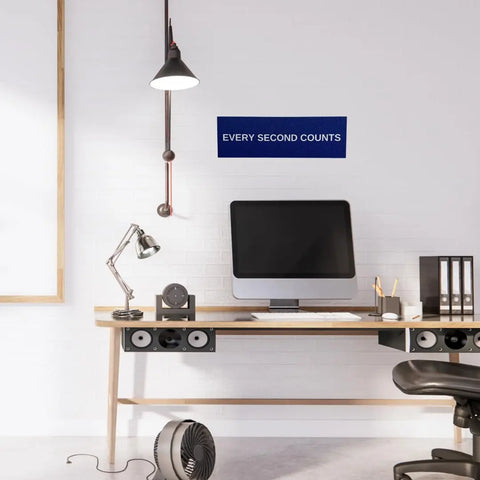 A modern desk setup with a computer, lamp, speakers, fan, and office supplies sits against a white brick wall. Above the desk, the Chatelet Manufacturing Every Second Counts Wall Sign adds an inspiring touch to the workspace.