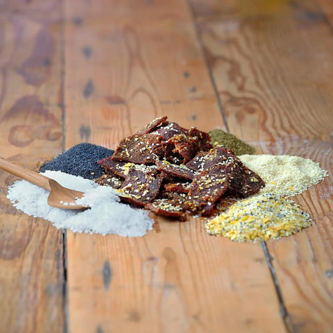 Field Trip Everything Bagel Beef Jerky 12oz is displayed on a wooden surface, surrounded by piles of coarse salt, seasoning, poppy seeds, dried herbs, and spices. A small wooden spoon rests in the salt beside the jerky.