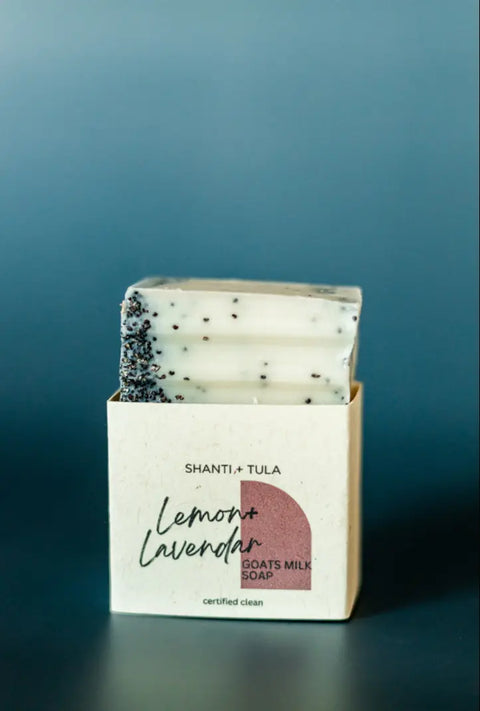 A Shanti + Tula Exfoliating Goat’s Milk Soap Bar with Pumice sits partly out of its box labeled Moisturizing & Handcrafted in Utah against a plain blue background. Ideal for exfoliating and moisturizing skincare routines.