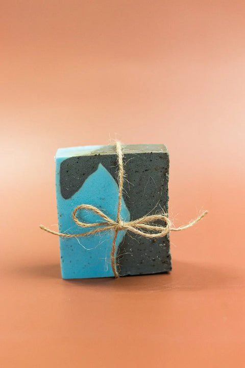 The Exfoliating Oatmeal & Shea Butter Body Scrub Bar by Shanti + Tula, handcrafted in the USA, features blue and dark gray hues, activated charcoal, and is tied with twine, displayed on a soft brown background.