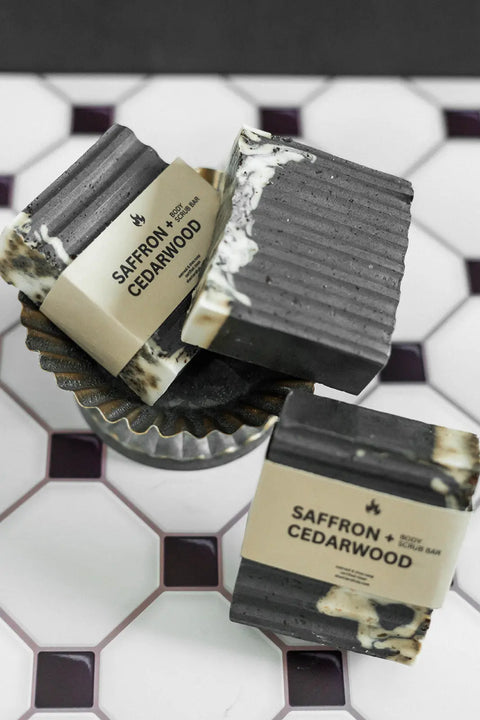 Three Exfoliating Oatmeal & Shea Butter Body Scrub Bars by Shanti + Tula rest on a geometric tile—two flat, one propped in a metal dish. These moisturizing, handcrafted-in-USA bars feature an elegant marbled look.