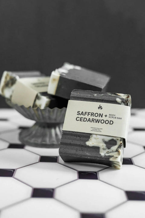 A Shanti + Tula Exfoliating Oatmeal & Shea Butter Body Scrub Bar, handcrafted in the USA, sits on geometric tile with more moisturizing bars stacked in a glass dish in the background.