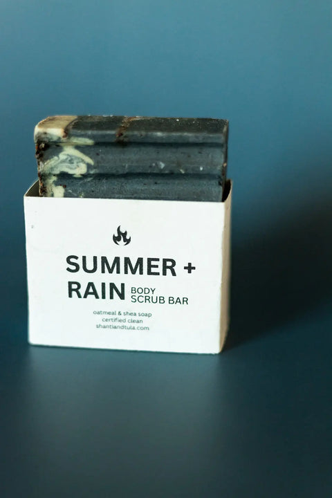 A dark, rectangular Shanti + Tula Exfoliating Oatmeal & Shea Butter Body Scrub Bar is partially inside a white box showing the product name, set against a blue background. The bar is moisturizing and handcrafted in the USA.